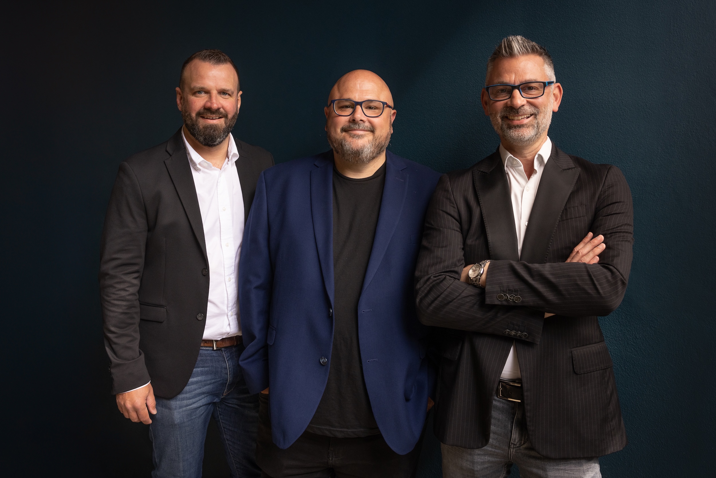 Three men in business attire pose for a professional photograph against a dark blue background. The man on the left wears a dark grey suit jacket with a white shirt and jeans. The man in the centre wears a blue suit jacket over a black shirt. The man on the right wears a dark pinstripe suit with a white shirt and has his arms crossed, wearing glasses and a watch.