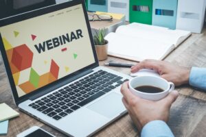 A person sat at a laptop with a cup of coffee. The word 'Webinar' is written on the laptop screen.