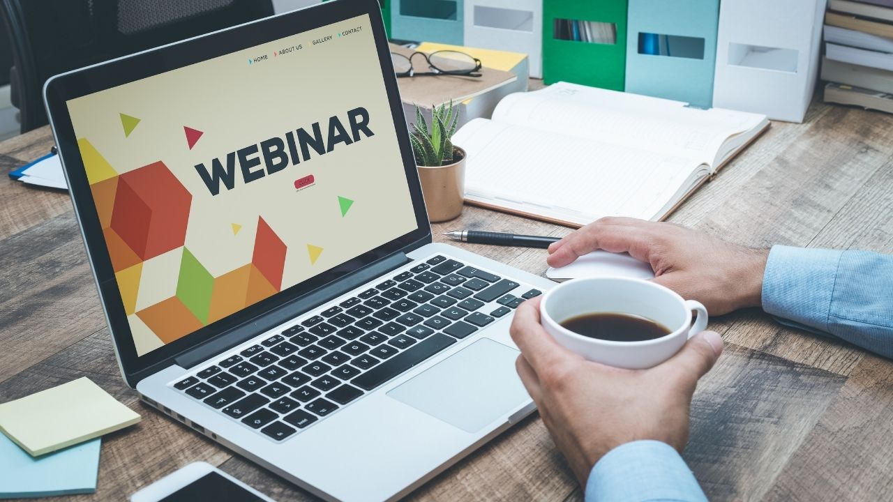 A person sat at a laptop with a cup of coffee. The word 'Webinar' is written on the laptop screen.