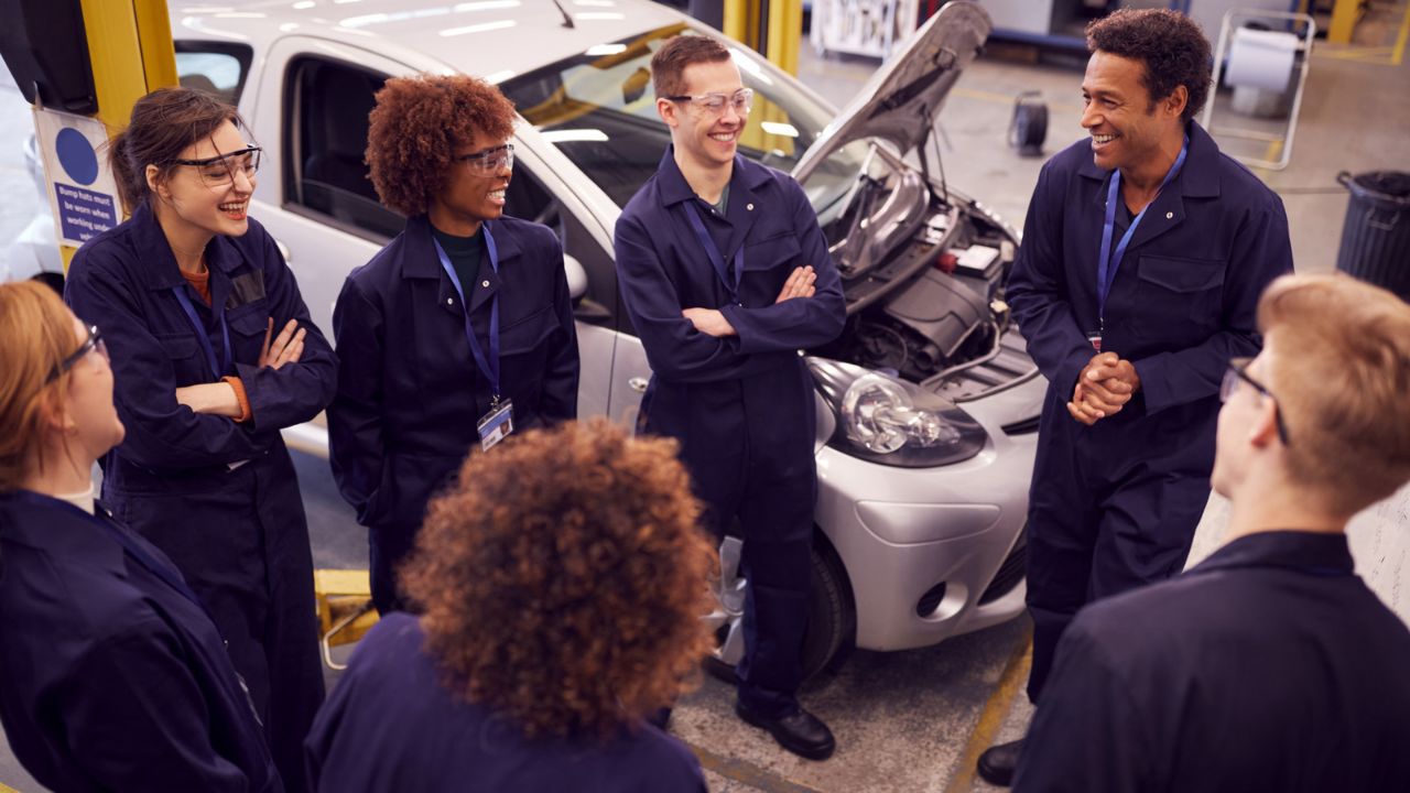 Six apprentices stood together with a mentor in a car garage.
